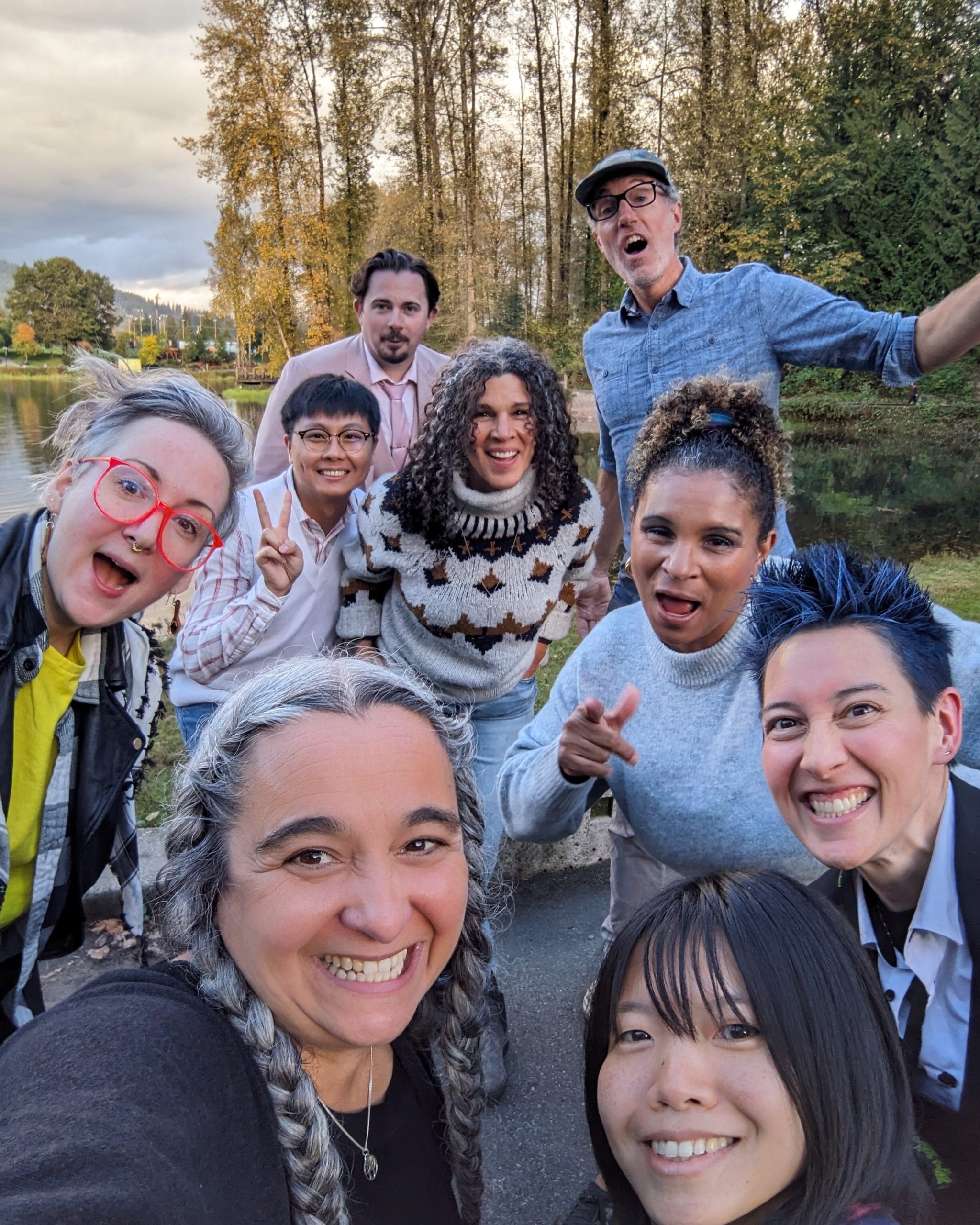 Pathways Team Photo A joyful group of ten adults poses for a selfie outdoors in front of a pond and tall trees. Everyone is smiling or making playful expressions. One person in front has long grey braids and is holding the camera. Others are making peace signs, pointing, or laughing, with the background showing autumn foliage and a partly cloudy sky.