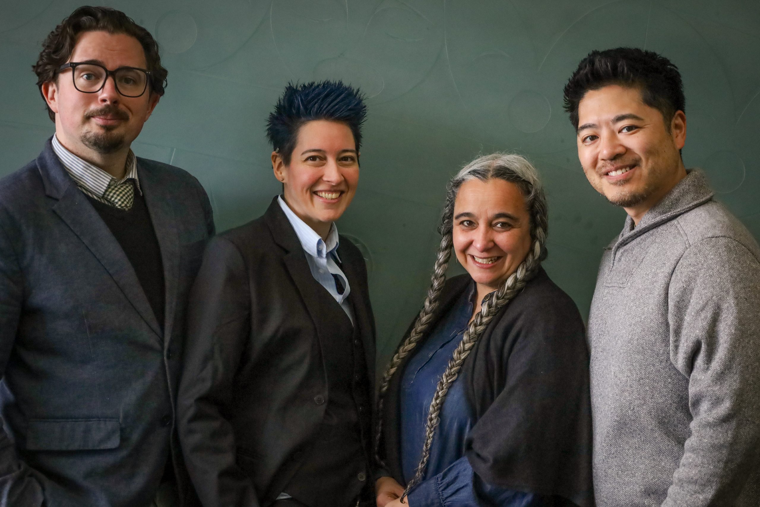 Co-Leadership Group Photo Four members of the leadership team stand in front of a green wall, smiling warmly at the camera. From left to right: Ryan has glasses and is wearing a dark suit and patterned tie; Elliott has short, spiked blue hair in a black suit and light blue shirt; Carla has long grey braids and is wearing a dark shawl over a navy blouse; and Kenji has short black hair and facial hair and is wearing a grey knit sweater. The setting appears professional and friendly.